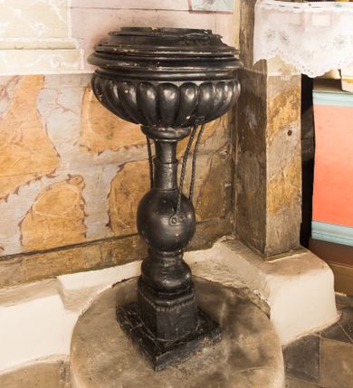 Zdjęcie nr 1: Chrzcielnica o kielichowym kształcie została ustawiona na dwustopniowej podstawie. Stopień pierwszy na rzucie koła jest kamienny, natomiast stopień drugi jest drewniany, prostopadłościenny, przechodzący w wieloboczną podstawę. Trzon chrzcielnicy uzyskał przekrój okrągły, w dolnej części wybrzuszony, a powyżej gruszkowaty. Czara jest spłaszczona, puklowana, zamknięta u góry profilowanym gzymsem. Przykrywa ją płaska nakrywa z profilowanymi bokami i metalowymi uchwytami. Dodatkowo czarę podtrzymują wtórnie dodane trzy pręty przymocowane do trzonu.