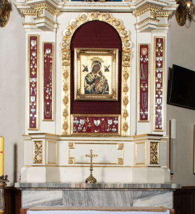 Zdjęcie nr 1: Ołtarz uzyskał formę architektonicznej struktury przyściennej, jednoosiowej, jednokondygnacyjnej ze zwieńczeniem. Marmurowy stipes ołtarzowy z mensą wykonano w formie prostopadłościanu dostawionego do wysokiego podwyższenia z marmuru ustawionego na podeście o ściętych narożach. Lico stipesu zdobi ryty symbol maryjny w otoku otoczonym przez stylizowane ramiona krzyża równoramiennego. Na podwyższeniu wsparto drewnianą strukturę nastawy ustawioną na cokole i flankowaną przez dwa filary. Przed lico kondygnacji wysunięto do przodu dwa postumenty, mające w pierwotnym zamyśle stanowić podpory dla flankujących strukturę dwóch kolumn podtrzymujący belkowanie wyłamane na ich osiach. Z kolumn zrezygnowano, a w ich miejsce w strukturę ołtarza oraz w trzony filarów zewnętrznych wprowadzono podłużne gabloty na wota. W polu kondygnacji wprowadzono prostokątną niszę zamkniętą półkoliście z mniejszym, czworobocznym obrazem Matki Boskiej Nieustającej Pomocy wiszącym na tle z szkarłatnej tkaniny. Poniżej obrazu umieszczono podłużną gablotę na wota. Ramę pola tworzą dwa pilastry o akantowych głowicach i trzonach dekorowanych zawieszonymi na taśmach bukiecikami kwiatów oraz półokrągły łuk, na którym umieszczono główki aniołków ze skrzyżowanymi skrzydełkami. Zwieńczenie utworzono z prostokątnego pola zamkniętego półkoliście ujętego obramieniem artykułowanym za pomocą rzędu kasetonów z rozetami, flankowanego przez dwa pilastry. W polu na niebieskim tle z tkaniny umieszczono ponad girlandą kwiatową w kształcie półksiężyca monogram maryjny także utworzony z girland roślino-kwiatowych. Zwieńczenie ujmuje para spływów wolutowych. Dwa pozostałe spływy wysunięte nad przedpolu zwieńczenia do przodu stanowią podstawę dla pary uskrzydlonych aniołków. Całość zwieńczenia zamyka kartusz prezentowany przez dwa aniołki oraz fantazyjny ornament spływający po jego bokach. W polu kartusza umieszczono napis: "O MARYJO / NIEUSTANNIE / WSPOMAGAJ / NAS". Struktura ołtarza polichromowana w kolorze kości słoniowej, profile i ornamenty zostały wyzłocone.  