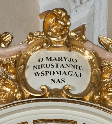 Zdjęcie nr 4: Ołtarz uzyskał formę architektonicznej struktury przyściennej, jednoosiowej, jednokondygnacyjnej ze zwieńczeniem. Marmurowy stipes ołtarzowy z mensą wykonano w formie prostopadłościanu dostawionego do wysokiego podwyższenia z marmuru ustawionego na podeście o ściętych narożach. Lico stipesu zdobi ryty symbol maryjny w otoku otoczonym przez stylizowane ramiona krzyża równoramiennego. Na podwyższeniu wsparto drewnianą strukturę nastawy ustawioną na cokole i flankowaną przez dwa filary. Przed lico kondygnacji wysunięto do przodu dwa postumenty, mające w pierwotnym zamyśle stanowić podpory dla flankujących strukturę dwóch kolumn podtrzymujący belkowanie wyłamane na ich osiach. Z kolumn zrezygnowano, a w ich miejsce w strukturę ołtarza oraz w trzony filarów zewnętrznych wprowadzono podłużne gabloty na wota. W polu kondygnacji wprowadzono prostokątną niszę zamkniętą półkoliście z mniejszym, czworobocznym obrazem Matki Boskiej Nieustającej Pomocy wiszącym na tle z szkarłatnej tkaniny. Poniżej obrazu umieszczono podłużną gablotę na wota. Ramę pola tworzą dwa pilastry o akantowych głowicach i trzonach dekorowanych zawieszonymi na taśmach bukiecikami kwiatów oraz półokrągły łuk, na którym umieszczono główki aniołków ze skrzyżowanymi skrzydełkami. Zwieńczenie utworzono z prostokątnego pola zamkniętego półkoliście ujętego obramieniem artykułowanym za pomocą rzędu kasetonów z rozetami, flankowanego przez dwa pilastry. W polu na niebieskim tle z tkaniny umieszczono ponad girlandą kwiatową w kształcie półksiężyca monogram maryjny także utworzony z girland roślino-kwiatowych. Zwieńczenie ujmuje para spływów wolutowych. Dwa pozostałe spływy wysunięte nad przedpolu zwieńczenia do przodu stanowią podstawę dla pary uskrzydlonych aniołków. Całość zwieńczenia zamyka kartusz prezentowany przez dwa aniołki oraz fantazyjny ornament spływający po jego bokach. W polu kartusza umieszczono napis: &quot;O MARYJO / NIEUSTANNIE / WSPOMAGAJ / NAS&quot;. Struktura ołtarza polichromowana w kolorze kości słoniowej, profile i ornamenty zostały wyzłocone.  