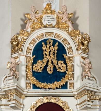 Zdjęcie nr 3: Ołtarz uzyskał formę architektonicznej struktury przyściennej, jednoosiowej, jednokondygnacyjnej ze zwieńczeniem. Marmurowy stipes ołtarzowy z mensą wykonano w formie prostopadłościanu dostawionego do wysokiego podwyższenia z marmuru ustawionego na podeście o ściętych narożach. Lico stipesu zdobi ryty symbol maryjny w otoku otoczonym przez stylizowane ramiona krzyża równoramiennego. Na podwyższeniu wsparto drewnianą strukturę nastawy ustawioną na cokole i flankowaną przez dwa filary. Przed lico kondygnacji wysunięto do przodu dwa postumenty, mające w pierwotnym zamyśle stanowić podpory dla flankujących strukturę dwóch kolumn podtrzymujący belkowanie wyłamane na ich osiach. Z kolumn zrezygnowano, a w ich miejsce w strukturę ołtarza oraz w trzony filarów zewnętrznych wprowadzono podłużne gabloty na wota. W polu kondygnacji wprowadzono prostokątną niszę zamkniętą półkoliście z mniejszym, czworobocznym obrazem Matki Boskiej Nieustającej Pomocy wiszącym na tle z szkarłatnej tkaniny. Poniżej obrazu umieszczono podłużną gablotę na wota. Ramę pola tworzą dwa pilastry o akantowych głowicach i trzonach dekorowanych zawieszonymi na taśmach bukiecikami kwiatów oraz półokrągły łuk, na którym umieszczono główki aniołków ze skrzyżowanymi skrzydełkami. Zwieńczenie utworzono z prostokątnego pola zamkniętego półkoliście ujętego obramieniem artykułowanym za pomocą rzędu kasetonów z rozetami, flankowanego przez dwa pilastry. W polu na niebieskim tle z tkaniny umieszczono ponad girlandą kwiatową w kształcie półksiężyca monogram maryjny także utworzony z girland roślino-kwiatowych. Zwieńczenie ujmuje para spływów wolutowych. Dwa pozostałe spływy wysunięte nad przedpolu zwieńczenia do przodu stanowią podstawę dla pary uskrzydlonych aniołków. Całość zwieńczenia zamyka kartusz prezentowany przez dwa aniołki oraz fantazyjny ornament spływający po jego bokach. W polu kartusza umieszczono napis: "O MARYJO / NIEUSTANNIE / WSPOMAGAJ / NAS". Struktura ołtarza polichromowana w kolorze kości słoniowej, profile i ornamenty zostały wyzłocone.  