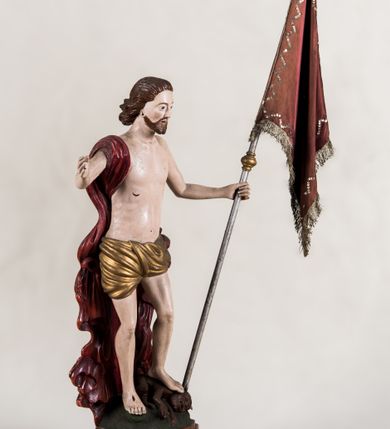 Zdjęcie nr 2: Pełnoplastyczna, całopostaciowa figura ukazuje Chrystusa Zmartwychwstałego stojącego na podłożu imitującym niewielki pagórek, osadzonym na profilowanej, prostopadłościennej podstawie o ściętych narożach. Zbawiciel na lekko ugiętych nogach ukazany został frontalnie. Obie ręce ma uniesione i odsunięte od tułowia. W lewej trzyma chorągiew na drzewcu z czerwonej tkaniny obszytej srebrną frędzlą i dekorowanej cekinami. Z kolei prawa jest uniesiona na wysokości klatki piersiowej. Jego sylwetka została opracowana sumarycznie z próbą uchwycenia muskulatury oraz zaznaczeniem partii żeber. Biodra Zbawiciela okrywa przewiązane, pozłocone perizonium. Chrystus ma owalną twarz o jasnej karnacji z charakterystycznie szeroko otwartymi oczami, wydatny nos i usta. Jego głowę otaczają krótkie brązowe włosy zaczesane do tyłu i zarost. Przez prawe ramię ma przerzucony, mocno sfałdowany, ciemnoczerwony płaszcz osłaniający tył sylwetki. Na dłoniach, stopach i w prawym boku widoczne są rany. Jezus lewą nogą depcze leżącego na podłożu szatana o groteskowej, brązowej sylwetce z ogonem, srebrzonymi, prostymi rogami, czerwonymi oczami oraz łapami zakończonymi czerwonymi pazurami. Na odwrocie widnieje napis: Odnowiono / 1960 St(Janisław) Rupik / Dzieło x(iędza) Roberta / Pioteckiego / 1615.
