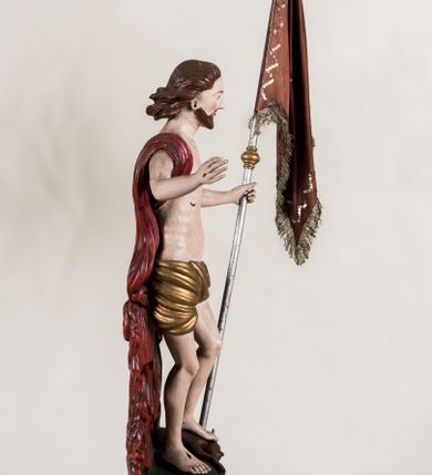 Zdjęcie nr 6: Pełnoplastyczna, całopostaciowa figura ukazuje Chrystusa Zmartwychwstałego stojącego na podłożu imitującym niewielki pagórek, osadzonym na profilowanej, prostopadłościennej podstawie o ściętych narożach. Zbawiciel na lekko ugiętych nogach ukazany został frontalnie. Obie ręce ma uniesione i odsunięte od tułowia. W lewej trzyma chorągiew na drzewcu z czerwonej tkaniny obszytej srebrną frędzlą i dekorowanej cekinami. Z kolei prawa jest uniesiona na wysokości klatki piersiowej. Jego sylwetka została opracowana sumarycznie z próbą uchwycenia muskulatury oraz zaznaczeniem partii żeber. Biodra Zbawiciela okrywa przewiązane, pozłocone perizonium. Chrystus ma owalną twarz o jasnej karnacji z charakterystycznie szeroko otwartymi oczami, wydatny nos i usta. Jego głowę otaczają krótkie brązowe włosy zaczesane do tyłu i zarost. Przez prawe ramię ma przerzucony, mocno sfałdowany, ciemnoczerwony płaszcz osłaniający tył sylwetki. Na dłoniach, stopach i w prawym boku widoczne są rany. Jezus lewą nogą depcze leżącego na podłożu szatana o groteskowej, brązowej sylwetce z ogonem, srebrzonymi, prostymi rogami, czerwonymi oczami oraz łapami zakończonymi czerwonymi pazurami. Na odwrocie widnieje napis: Odnowiono / 1960 St(Janisław) Rupik / Dzieło x(iędza) Roberta / Pioteckiego / 1615.
