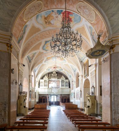 Zdjęcie nr 18: Po kilkukrotnych przekształceniach kościół składa się z prostokątnej, czteroprzęsłowej nawy głównej, dwóch, dwuprzęsłowych naw bocznych, przerobionych z dawnych kaplic i jednoprzęsłowego prezbiterium zamkniętego od wschodu apsydą. Na przedłużeniu apsydy otoczonej półkolistym aneksem wzniesiono czworoboczną, piętrową zakrystią. Od zachodu do nawy przylega wysunięta do przodu czworoboczna wieża o ściętych narożach z urządzoną w przyziemiu kruchtą. Wieża uzyskała trzy kondygnacje wyznaczone za pomocą profilowanych gzymsów. Środkowe piętro wieży połączono z bocznymi częściami fasady za pomocą wolut. Na dwóch pierwszych kondygnacjach ścięte naroża artykułowano za pomocą pilastrów o prostych głowicach. Pilastry drugiej kondygnacji ozdobiono krótkimi zwisami kampanul. Kondygnacja trzecia także została wzniesiona na rzucie czworoboku ale o wklęsłych narożach. Jej ściany flankują pary pilastrów o korynckich głowicach podtrzymujące imposty wspierające półkoliście wyłamane na osi belkowanie wspierające baniasty hełm z latarnią zwieńczony krzyżem z koroną. Pomiędzy impostami pod wyłamanym belkowaniem umieszczono tarcze zegarowe. Na trzeciej kondygnacji ponad belkowaniem od zachodu nadwieszono czworoboczny balkon z tralkową balustradą. Sześcioboczną sygnaturkę z latarnią zwieńczoną stożkowym hełmem z ustawioną na szczycie figurką Matki Boskiej Niepokalanie Poczętej zawieszono na siodłowym dachu kościoła w części wschodniej nawy głównej. Elewacje boczne kościoła rozczłonkowano za pomocą podziałów ramowych lub lizenowych. Wnętrze świątyni nakryto sklepieniem kolebkowym wspartym na gurtach, przechodzących na przyścienne pilastry podtrzymujące odcinki przerwanego belkowania. W ostanim zachodnim przęśle urządzono chór muzyczny wsparty na dwóch filarach z wypukłym na osi parapetem, artykułowanym za pomocą pilastrów i par arkadek. W prezbiterium ustawiono ołtarz główny oraz ambonę, a w nawach ołtarze boczne. 