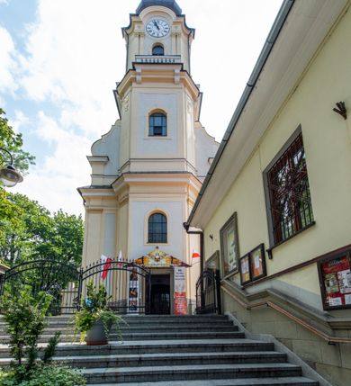 Zdjęcie nr 27: Po kilkukrotnych przekształceniach kościół składa się z prostokątnej, czteroprzęsłowej nawy głównej, dwóch, dwuprzęsłowych naw bocznych, przerobionych z dawnych kaplic i jednoprzęsłowego prezbiterium zamkniętego od wschodu apsydą. Na przedłużeniu apsydy otoczonej półkolistym aneksem wzniesiono czworoboczną, piętrową zakrystią. Od zachodu do nawy przylega wysunięta do przodu czworoboczna wieża o ściętych narożach z urządzoną w przyziemiu kruchtą. Wieża uzyskała trzy kondygnacje wyznaczone za pomocą profilowanych gzymsów. Środkowe piętro wieży połączono z bocznymi częściami fasady za pomocą wolut. Na dwóch pierwszych kondygnacjach ścięte naroża artykułowano za pomocą pilastrów o prostych głowicach. Pilastry drugiej kondygnacji ozdobiono krótkimi zwisami kampanul. Kondygnacja trzecia także została wzniesiona na rzucie czworoboku ale o wklęsłych narożach. Jej ściany flankują pary pilastrów o korynckich głowicach podtrzymujące imposty wspierające półkoliście wyłamane na osi belkowanie wspierające baniasty hełm z latarnią zwieńczony krzyżem z koroną. Pomiędzy impostami pod wyłamanym belkowaniem umieszczono tarcze zegarowe. Na trzeciej kondygnacji ponad belkowaniem od zachodu nadwieszono czworoboczny balkon z tralkową balustradą. Sześcioboczną sygnaturkę z latarnią zwieńczoną stożkowym hełmem z ustawioną na szczycie figurką Matki Boskiej Niepokalanie Poczętej zawieszono na siodłowym dachu kościoła w części wschodniej nawy głównej. Elewacje boczne kościoła rozczłonkowano za pomocą podziałów ramowych lub lizenowych. Wnętrze świątyni nakryto sklepieniem kolebkowym wspartym na gurtach, przechodzących na przyścienne pilastry podtrzymujące odcinki przerwanego belkowania. W ostanim zachodnim przęśle urządzono chór muzyczny wsparty na dwóch filarach z wypukłym na osi parapetem, artykułowanym za pomocą pilastrów i par arkadek. W prezbiterium ustawiono ołtarz główny oraz ambonę, a w nawach ołtarze boczne. 