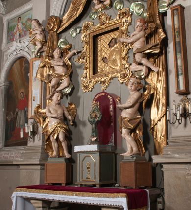 Zdjęcie nr 44: Nastawa niearchitektoniczna w formie paludamentu. Mensa kamienna, oparta na dwóch kamiennych pilastrach o kanelowanych trzonach, ustawionych na cokolikach. Na mensie  tabernakulum w formie drewnianej skrzynki na czterech nóżkach, zakończone gzymsem, o drzwiczkach w kształcie stojącego prostokąta o ściętych górnych narożach, ujęte profilowaną ramą z przedstawieniem kielicha z hostią w glorii. Na nim tron wystawienia na relikwiarz. Tabernakulum flankują dwie figury aniołów ustawione na cokołach. 
Paludament podwiązany po bokach sznurem z chwostami, podtrzymywany przez putta, lamowany frędzlą, zwieńczony baldachimem utworzonym z fragmentu wklęsło-wypukłego gzymsu z lambrekinem. W polu wnęka w kształcie stojącego prostokąta ujęta profilowaną ramą i rocaille'm, z obrazem św. Karola Boromeusza. Nad obrazem rzeźbione obłoki i dwie uskrzydlone główki anielskie, po bokach nadlatujące dwa putta. Paludament polichromowany wewnątrz w kolorze szaro-brązowym, zewnątrz złocony.