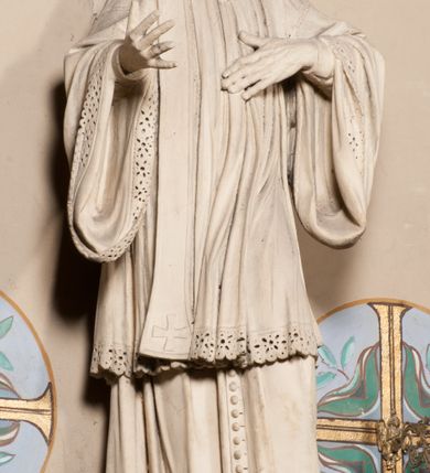 Zdjęcie nr 8: Pełnoplastyczna rzeźba ustawiona na czworobocznym, niskim postumencie wspartym na przyściennej, wolutowej konsoli przedstawia w całej postaci ujętego w nieznacznym kontrapoście księdza. Duchowny prawą rękę ma odsuniętą od siebie, lewą zaś przykłada do piersi. Jest to młody mężczyzna z uniesioną głową i spojrzeniem skierowanym w niebo. Ma owalną twarz z wydatnym nosem i ustami pozbawioną zarostu oraz krótkie włosy odsłaniające uszy. Duchowny ubrany jest w sutannę, z wysoką koloratką, komżę wiązaną pod szyją z szerokimi rękawami, obszytą koronką oraz nałożoną na ramionach stułę. Spod sutanny widać trzewiki. Rzeźba w całości polichromowana jest na biało. Na postumencie wyryta data 1797. 