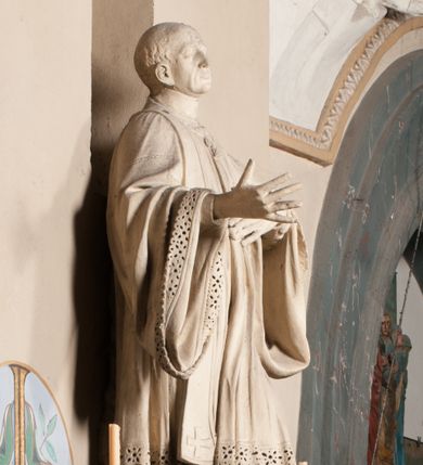 Zdjęcie nr 1: Pełnoplastyczna rzeźba ustawiona na czworobocznym, niskim postumencie wspartym na przyściennej, wolutowej konsoli przedstawia w całej postaci ujętego w nieznacznym kontrapoście księdza. Duchowny prawą rękę ma odsuniętą od siebie, lewą zaś przykłada do piersi. Jest to młody mężczyzna z uniesioną głową i spojrzeniem skierowanym w niebo. Ma owalną twarz z wydatnym nosem i ustami pozbawioną zarostu oraz krótkie włosy odsłaniające uszy. Duchowny ubrany jest w sutannę, z wysoką koloratką, komżę wiązaną pod szyją z szerokimi rękawami, obszytą koronką oraz nałożoną na ramionach stułę. Spod sutanny widać trzewiki. Rzeźba w całości polichromowana jest na biało. Na postumencie wyryta data 1797. 