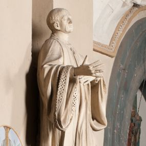 Zdjęcie nr 1: Pełnoplastyczna rzeźba ustawiona na czworobocznym, niskim postumencie wspartym na przyściennej, wolutowej konsoli przedstawia w całej postaci ujętego w nieznacznym kontrapoście księdza. Duchowny prawą rękę ma odsuniętą od siebie, lewą zaś przykłada do piersi. Jest to młody mężczyzna z uniesioną głową i spojrzeniem skierowanym w niebo. Ma owalną twarz z wydatnym nosem i ustami pozbawioną zarostu oraz krótkie włosy odsłaniające uszy. Duchowny ubrany jest w sutannę, z wysoką koloratką, komżę wiązaną pod szyją z szerokimi rękawami, obszytą koronką oraz nałożoną na ramionach stułę. Spod sutanny widać trzewiki. Rzeźba w całości polichromowana jest na biało. Na postumencie wyryta data 1797. 