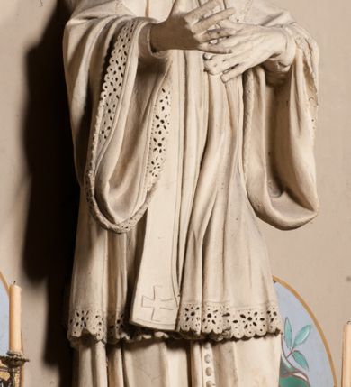 Zdjęcie nr 7: Pełnoplastyczna rzeźba ustawiona na czworobocznym, niskim postumencie wspartym na przyściennej, wolutowej konsoli przedstawia w całej postaci ujętego w nieznacznym kontrapoście księdza. Duchowny prawą rękę ma odsuniętą od siebie, lewą zaś przykłada do piersi. Jest to młody mężczyzna z uniesioną głową i spojrzeniem skierowanym w niebo. Ma owalną twarz z wydatnym nosem i ustami pozbawioną zarostu oraz krótkie włosy odsłaniające uszy. Duchowny ubrany jest w sutannę, z wysoką koloratką, komżę wiązaną pod szyją z szerokimi rękawami, obszytą koronką oraz nałożoną na ramionach stułę. Spod sutanny widać trzewiki. Rzeźba w całości polichromowana jest na biało. Na postumencie wyryta data 1797. 