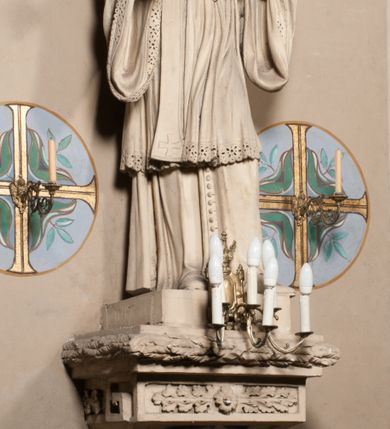 Zdjęcie nr 2: Pełnoplastyczna rzeźba ustawiona na czworobocznym, niskim postumencie wspartym na przyściennej, wolutowej konsoli przedstawia w całej postaci ujętego w nieznacznym kontrapoście księdza. Duchowny prawą rękę ma odsuniętą od siebie, lewą zaś przykłada do piersi. Jest to młody mężczyzna z uniesioną głową i spojrzeniem skierowanym w niebo. Ma owalną twarz z wydatnym nosem i ustami pozbawioną zarostu oraz krótkie włosy odsłaniające uszy. Duchowny ubrany jest w sutannę, z wysoką koloratką, komżę wiązaną pod szyją z szerokimi rękawami, obszytą koronką oraz nałożoną na ramionach stułę. Spod sutanny widać trzewiki. Rzeźba w całości polichromowana jest na biało. Na postumencie wyryta data 1797. 