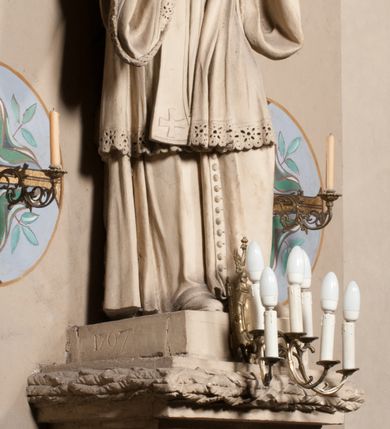 Zdjęcie nr 9: Pełnoplastyczna rzeźba ustawiona na czworobocznym, niskim postumencie wspartym na przyściennej, wolutowej konsoli przedstawia w całej postaci ujętego w nieznacznym kontrapoście księdza. Duchowny prawą rękę ma odsuniętą od siebie, lewą zaś przykłada do piersi. Jest to młody mężczyzna z uniesioną głową i spojrzeniem skierowanym w niebo. Ma owalną twarz z wydatnym nosem i ustami pozbawioną zarostu oraz krótkie włosy odsłaniające uszy. Duchowny ubrany jest w sutannę, z wysoką koloratką, komżę wiązaną pod szyją z szerokimi rękawami, obszytą koronką oraz nałożoną na ramionach stułę. Spod sutanny widać trzewiki. Rzeźba w całości polichromowana jest na biało. Na postumencie wyryta data 1797. 