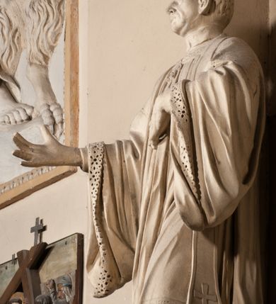 Zdjęcie nr 14: Pełnoplastyczna rzeźba ustawiona na czworobocznym, niskim postumencie wspartym na przyściennej, wolutowej konsoli przedstawia w całej postaci ujętego w nieznacznym kontrapoście księdza. Duchowny prawą rękę ma odsuniętą od siebie, lewą zaś przykłada do piersi. Jest to młody mężczyzna z uniesioną głową i spojrzeniem skierowanym w niebo. Ma owalną twarz z wydatnym nosem i ustami pozbawioną zarostu oraz krótkie włosy odsłaniające uszy. Duchowny ubrany jest w sutannę, z wysoką koloratką, komżę wiązaną pod szyją z szerokimi rękawami, obszytą koronką oraz nałożoną na ramionach stułę. Spod sutanny widać trzewiki. Rzeźba w całości polichromowana jest na biało. Na postumencie wyryta data 1797. 