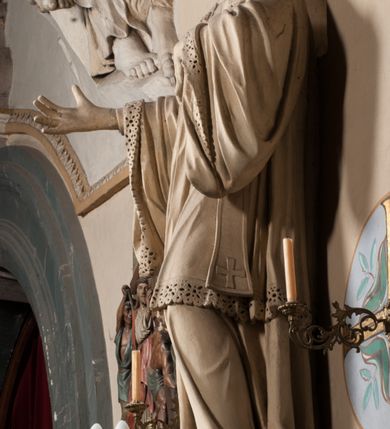 Zdjęcie nr 3: Pełnoplastyczna rzeźba ustawiona na czworobocznym, niskim postumencie wspartym na przyściennej, wolutowej konsoli przedstawia w całej postaci ujętego w nieznacznym kontrapoście księdza. Duchowny prawą rękę ma odsuniętą od siebie, lewą zaś przykłada do piersi. Jest to młody mężczyzna z uniesioną głową i spojrzeniem skierowanym w niebo. Ma owalną twarz z wydatnym nosem i ustami pozbawioną zarostu oraz krótkie włosy odsłaniające uszy. Duchowny ubrany jest w sutannę, z wysoką koloratką, komżę wiązaną pod szyją z szerokimi rękawami, obszytą koronką oraz nałożoną na ramionach stułę. Spod sutanny widać trzewiki. Rzeźba w całości polichromowana jest na biało. Na postumencie wyryta data 1797. 