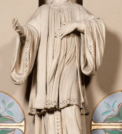 Zdjęcie nr 6: Pełnoplastyczna rzeźba ustawiona na czworobocznym, niskim postumencie wspartym na przyściennej, wolutowej konsoli przedstawia w całej postaci ujętego w nieznacznym kontrapoście księdza. Duchowny prawą rękę ma odsuniętą od siebie, lewą zaś przykłada do piersi. Jest to młody mężczyzna z uniesioną głową i spojrzeniem skierowanym w niebo. Ma owalną twarz z wydatnym nosem i ustami pozbawioną zarostu oraz krótkie włosy odsłaniające uszy. Duchowny ubrany jest w sutannę, z wysoką koloratką, komżę wiązaną pod szyją z szerokimi rękawami, obszytą koronką oraz nałożoną na ramionach stułę. Spod sutanny widać trzewiki. Rzeźba w całości polichromowana jest na biało. Na postumencie wyryta data 1797. 