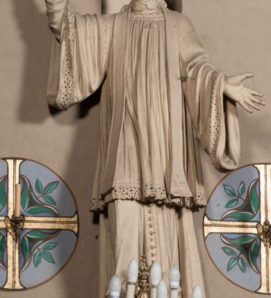 Zdjęcie nr 4: Pełnoplastyczna rzeźba ustawiona na czworobocznym, niskim postumencie wspartym na przyściennej, wolutowej konsoli przedstawia w całej postaci ujętego w nieznacznym kontrapoście księdza. Duchowny prawą rękę wznosi ku górze, lewą zaś odsuwa od siebie. Jest to młody mężczyzna z uniesioną głową i spojrzeniem skierowanym w niebo. Ma owalną twarz z wydatnym nosem i ustami okoloną krótkim zarostem i zmierzwionymi włosami odsłaniającymi uszy oraz spływającymi na ramiona. Duchowny ubrany jest w sutannę, z wysoką koloratką, komżę wiązaną pod szyją z szerokimi rękawami obszytą koronką oraz nałożoną na ramionach stułę. Spod sutanny widać trzewiki. Rzeźba w całości polichromowana jest na biało.