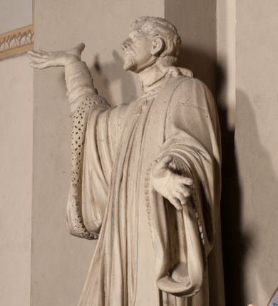 Zdjęcie nr 1: Pełnoplastyczna rzeźba ustawiona na czworobocznym, niskim postumencie wspartym na przyściennej, wolutowej konsoli przedstawia w całej postaci ujętego w nieznacznym kontrapoście księdza. Duchowny prawą rękę wznosi ku górze, lewą zaś odsuwa od siebie. Jest to młody mężczyzna z uniesioną głową i spojrzeniem skierowanym w niebo. Ma owalną twarz z wydatnym nosem i ustami okoloną krótkim zarostem i zmierzwionymi włosami odsłaniającymi uszy oraz spływającymi na ramiona. Duchowny ubrany jest w sutannę, z wysoką koloratką, komżę wiązaną pod szyją z szerokimi rękawami obszytą koronką oraz nałożoną na ramionach stułę. Spod sutanny widać trzewiki. Rzeźba w całości polichromowana jest na biało.