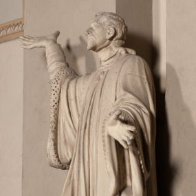 Zdjęcie nr 1: Pełnoplastyczna rzeźba ustawiona na czworobocznym, niskim postumencie wspartym na przyściennej, wolutowej konsoli przedstawia w całej postaci ujętego w nieznacznym kontrapoście księdza. Duchowny prawą rękę wznosi ku górze, lewą zaś odsuwa od siebie. Jest to młody mężczyzna z uniesioną głową i spojrzeniem skierowanym w niebo. Ma owalną twarz z wydatnym nosem i ustami okoloną krótkim zarostem i zmierzwionymi włosami odsłaniającymi uszy oraz spływającymi na ramiona. Duchowny ubrany jest w sutannę, z wysoką koloratką, komżę wiązaną pod szyją z szerokimi rękawami obszytą koronką oraz nałożoną na ramionach stułę. Spod sutanny widać trzewiki. Rzeźba w całości polichromowana jest na biało.