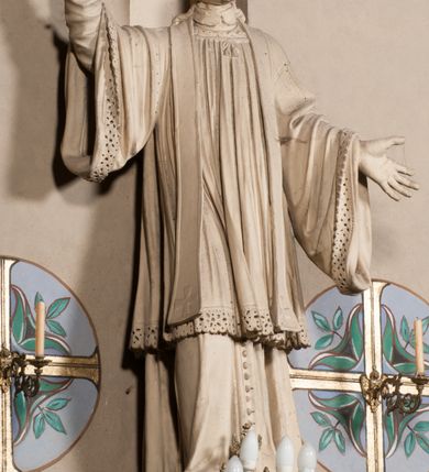 Zdjęcie nr 2: Pełnoplastyczna rzeźba ustawiona na czworobocznym, niskim postumencie wspartym na przyściennej, wolutowej konsoli przedstawia w całej postaci ujętego w nieznacznym kontrapoście księdza. Duchowny prawą rękę wznosi ku górze, lewą zaś odsuwa od siebie. Jest to młody mężczyzna z uniesioną głową i spojrzeniem skierowanym w niebo. Ma owalną twarz z wydatnym nosem i ustami okoloną krótkim zarostem i zmierzwionymi włosami odsłaniającymi uszy oraz spływającymi na ramiona. Duchowny ubrany jest w sutannę, z wysoką koloratką, komżę wiązaną pod szyją z szerokimi rękawami obszytą koronką oraz nałożoną na ramionach stułę. Spod sutanny widać trzewiki. Rzeźba w całości polichromowana jest na biało.