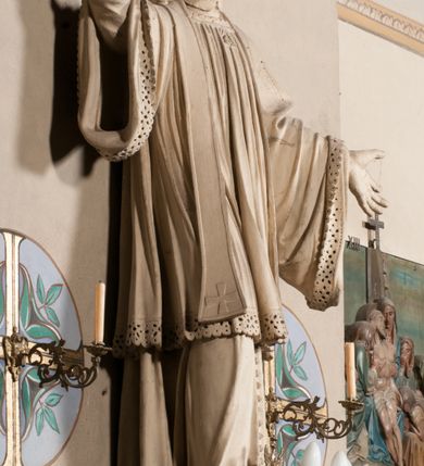 Zdjęcie nr 3: Pełnoplastyczna rzeźba ustawiona na czworobocznym, niskim postumencie wspartym na przyściennej, wolutowej konsoli przedstawia w całej postaci ujętego w nieznacznym kontrapoście księdza. Duchowny prawą rękę wznosi ku górze, lewą zaś odsuwa od siebie. Jest to młody mężczyzna z uniesioną głową i spojrzeniem skierowanym w niebo. Ma owalną twarz z wydatnym nosem i ustami okoloną krótkim zarostem i zmierzwionymi włosami odsłaniającymi uszy oraz spływającymi na ramiona. Duchowny ubrany jest w sutannę, z wysoką koloratką, komżę wiązaną pod szyją z szerokimi rękawami obszytą koronką oraz nałożoną na ramionach stułę. Spod sutanny widać trzewiki. Rzeźba w całości polichromowana jest na biało.