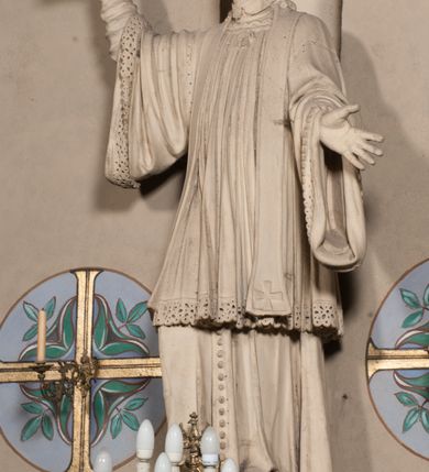 Zdjęcie nr 5: Pełnoplastyczna rzeźba ustawiona na czworobocznym, niskim postumencie wspartym na przyściennej, wolutowej konsoli przedstawia w całej postaci ujętego w nieznacznym kontrapoście księdza. Duchowny prawą rękę wznosi ku górze, lewą zaś odsuwa od siebie. Jest to młody mężczyzna z uniesioną głową i spojrzeniem skierowanym w niebo. Ma owalną twarz z wydatnym nosem i ustami okoloną krótkim zarostem i zmierzwionymi włosami odsłaniającymi uszy oraz spływającymi na ramiona. Duchowny ubrany jest w sutannę, z wysoką koloratką, komżę wiązaną pod szyją z szerokimi rękawami obszytą koronką oraz nałożoną na ramionach stułę. Spod sutanny widać trzewiki. Rzeźba w całości polichromowana jest na biało.