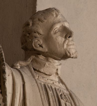 Zdjęcie nr 6: Pełnoplastyczna rzeźba ustawiona na czworobocznym, niskim postumencie wspartym na przyściennej, wolutowej konsoli przedstawia w całej postaci ujętego w nieznacznym kontrapoście księdza. Duchowny prawą rękę wznosi ku górze, lewą zaś odsuwa od siebie. Jest to młody mężczyzna z uniesioną głową i spojrzeniem skierowanym w niebo. Ma owalną twarz z wydatnym nosem i ustami okoloną krótkim zarostem i zmierzwionymi włosami odsłaniającymi uszy oraz spływającymi na ramiona. Duchowny ubrany jest w sutannę, z wysoką koloratką, komżę wiązaną pod szyją z szerokimi rękawami obszytą koronką oraz nałożoną na ramionach stułę. Spod sutanny widać trzewiki. Rzeźba w całości polichromowana jest na biało.