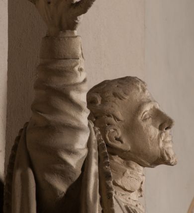 Zdjęcie nr 9: Pełnoplastyczna rzeźba ustawiona na czworobocznym, niskim postumencie wspartym na przyściennej, wolutowej konsoli przedstawia w całej postaci ujętego w nieznacznym kontrapoście księdza. Duchowny prawą rękę wznosi ku górze, lewą zaś odsuwa od siebie. Jest to młody mężczyzna z uniesioną głową i spojrzeniem skierowanym w niebo. Ma owalną twarz z wydatnym nosem i ustami okoloną krótkim zarostem i zmierzwionymi włosami odsłaniającymi uszy oraz spływającymi na ramiona. Duchowny ubrany jest w sutannę, z wysoką koloratką, komżę wiązaną pod szyją z szerokimi rękawami obszytą koronką oraz nałożoną na ramionach stułę. Spod sutanny widać trzewiki. Rzeźba w całości polichromowana jest na biało.