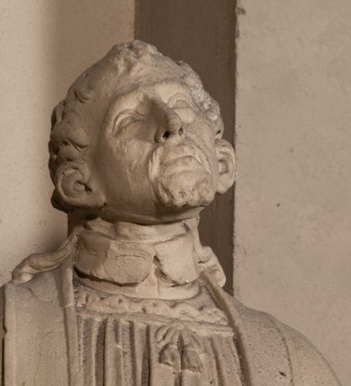 Zdjęcie nr 7: Pełnoplastyczna rzeźba ustawiona na czworobocznym, niskim postumencie wspartym na przyściennej, wolutowej konsoli przedstawia w całej postaci ujętego w nieznacznym kontrapoście księdza. Duchowny prawą rękę wznosi ku górze, lewą zaś odsuwa od siebie. Jest to młody mężczyzna z uniesioną głową i spojrzeniem skierowanym w niebo. Ma owalną twarz z wydatnym nosem i ustami okoloną krótkim zarostem i zmierzwionymi włosami odsłaniającymi uszy oraz spływającymi na ramiona. Duchowny ubrany jest w sutannę, z wysoką koloratką, komżę wiązaną pod szyją z szerokimi rękawami obszytą koronką oraz nałożoną na ramionach stułę. Spod sutanny widać trzewiki. Rzeźba w całości polichromowana jest na biało.