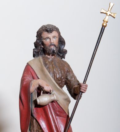 Zdjęcie nr 1: Rzeźba ustawiona na pokrywie chrzcielnicy, pełna, przedstawiająca św. Jana Chrzciciela. Święty ukazany w całej postaci, w delikatnym kontrapoście. W prawej dłoni trzyma dzbanuszek do polania wodą, a w lewej równoramienny krzyż na długim drzewcu. U jego prawej nogi leży biały baranek. Twarz podłużna o sumarycznie oddanych detalach, małe oczy i usta, krótki nos. Włosy bujne, ciemnobrązowe, opadają na plecy i ramiona, broda krótka rozdzielona na końcu w dwa pukle. Święty ubrany jest w suknię wykonaną z wielbłądziej skóry, sięgającą połowy łydek oraz czerwony płaszcz z jasnobeżową podszewką założony na prawe ramię i spływający pod lewą ręką. Draperia szat spokojna, niewielka. Polichromia w odsłoniętych partiach ciała naturalistyczna. 