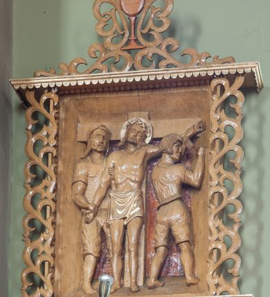 Zdjęcie nr 3: Stacje drogi krzyżowej w formie płaskorzeźbionych scen ujętych rozbudowanym obramieniem. Przedstawienia ograniczają się do najważniejszych osób, ukazanych w całej postaci i bliskim planie, na jednolitym, nieco ciemniejszym od reszty tle. Sposób ukazania postaci nosi cechy sztuki ludowej, ich pozy są sztywne, twarze zwrócone do widza, szaty marszczą się drobnymi fałdami. Postacie, ich ubrania oraz elementy scen zachowują naturalny kolor drewna, jedynie ubrania i nimb Chrystusa podkreślono złoceniem. Sceny zakomponowano w obramienie przypominające kapliczkę – na podstawie i pod daszkiem dekorowanymi lambrekinem, ujęte po bokach ażurową wicią roślinną. Poniżej podstawy numer sceny, a powyżej kielich z zatkniętym krzyżem łacińskim otacza i łączy w całość podobna wić. Lambrekin, krzyż oraz cyfry rzymskie złocone, reszta w kolorze naturalnego drewna. 