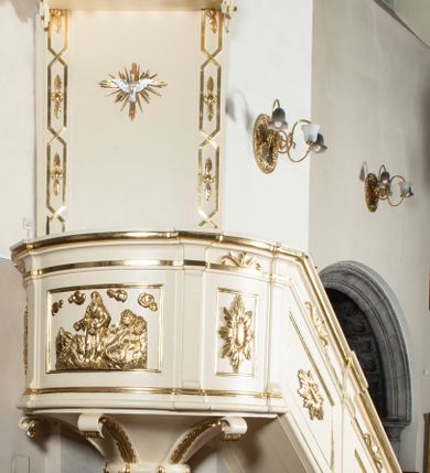 Zdjęcie nr 1: Przyścienna, wisząca z dostępem w formie jednobiegowych schodów od strony prezbiterium, ambona składa się z kosza, zaplecka i baldachimu. Kosz został wykonany na planie półkola ustawionego na niskim cokole z podwieszeniem w formie zbliżonej do stożka, ujętego wolutami i zakończonego wałkiem z szyszką. Jego boki podzielono pilastrami na nierówne pola, w których znajdują się płyciny z rozetami w zewnętrznych polach oraz w środkowym płaskorzeźbione przedstawienie Chrystusa siewcy na tle krajobrazu siewcy. Kosz zwieńczony został parapetem w formie gzymsu. Zaplecek ambony uzyskał formę stojącego prostokąta, ujętego lizenami ozdobionymi dekoracją geometryczną i roślinną. W polu umieszczono płaskorzeźbioną gołębicę Ducha Świętego w glorii promienistej. Baldachim wykonano na planie półkola w formie belkowania z podwieszonym lambrekinem i fryzem dekorowanym diagonalną kratownicą. Górę baldachimu opięto belkowaniem wyłamanym na dwóch osiach skrajnych. Baldachim wieńczy postument z leżącą na nim zamkniętą księgą z barankiem apokaliptycznym, stojącymi za nim dwiema tablicami Dekalogu oraz widniejącym nad nimi krzyżem w promienistej glorii. Architektura ambony została polichromowana na biało ze złoconymi profilami i ornamentyką oraz srebrzonymi detalami.