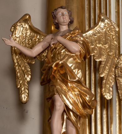 Zdjęcie nr 1: Rzeźba anioła jest ścięta i drążona z tyłu. Ustawiono ją na prostokątnej niskiej podstawie. Anioł ukazany został frontalnie w kontrapoście. Lewą rękę kładzie na piersi, a prawą wyciąga w bok. Ma owalną twarz o szeroko otwartych oczach, małym nosie, pełnych ustach, okoloną krótkimi, silnie kręconymi włosami. Ubrany jest w tunikę odsłaniającą prawe ramię, z rękawem na lewym ramieniu, przewiązaną w talii, odsłaniającą prawe kolano oraz płaszcz przerzucony przez lewe ramię, otaczający postać, podtrzymany z przodu na tasiemce. Bo bokach widoczne są złożone skrzydła. Rzeźba jest naturalistycznie polichromowana, a szaty i skrzydła zostały wyzłocone.