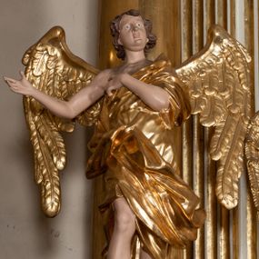 Zdjęcie nr 1: Rzeźba anioła jest ścięta i drążona z tyłu. Ustawiono ją na prostokątnej niskiej podstawie. Anioł ukazany został frontalnie w kontrapoście. Lewą rękę kładzie na piersi, a prawą wyciąga w bok. Ma owalną twarz o szeroko otwartych oczach, małym nosie, pełnych ustach, okoloną krótkimi, silnie kręconymi włosami. Ubrany jest w tunikę odsłaniającą prawe ramię, z rękawem na lewym ramieniu, przewiązaną w talii, odsłaniającą prawe kolano oraz płaszcz przerzucony przez lewe ramię, otaczający postać, podtrzymany z przodu na tasiemce. Bo bokach widoczne są złożone skrzydła. Rzeźba jest naturalistycznie polichromowana, a szaty i skrzydła zostały wyzłocone.