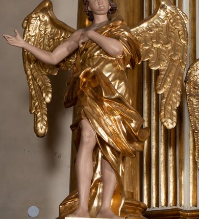 Zdjęcie nr 2: Rzeźba anioła jest ścięta i drążona z tyłu. Ustawiono ją na prostokątnej niskiej podstawie. Anioł ukazany został frontalnie w kontrapoście. Lewą rękę kładzie na piersi, a prawą wyciąga w bok. Ma owalną twarz o szeroko otwartych oczach, małym nosie, pełnych ustach, okoloną krótkimi, silnie kręconymi włosami. Ubrany jest w tunikę odsłaniającą prawe ramię, z rękawem na lewym ramieniu, przewiązaną w talii, odsłaniającą prawe kolano oraz płaszcz przerzucony przez lewe ramię, otaczający postać, podtrzymany z przodu na tasiemce. Bo bokach widoczne są złożone skrzydła. Rzeźba jest naturalistycznie polichromowana, a szaty i skrzydła zostały wyzłocone.