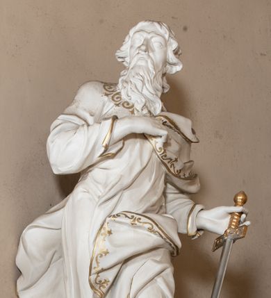 Zdjęcie nr 1: Rzeźba drążona z tyłu stoi na prostokątnej podstawie ustawionej na wolutowej konsoli przyściennej. Postać apostoła jest ujęta frontalnie w kontrapoście z głową skierowaną w prawo. Prawą rękę Paweł ma złożoną na piersi, lewą zaś wyciągniętą w bok trzyma miecz zwrócony ostrzem w dół. Święty ma pociągłą, wychudzoną twarz o migdałowatych oczach, lekko garbatym nosie i pełnych rozchylonych ustach. Głowę okala opadająca na pierś broda i lekko kręcone sięgające ramion włosy. Postać ubrana jest w długą, przylegającą do ciała suknię oraz płaszcz zarzucony na lewę ramię, opadający na plecy z fragmentem tkaniny przerzuconym przez lewe biodro. Rzeźba jest polichromowana w całości na biało ze złoconymi lamówkami szat i wzorem dekoracyjnym wzdłuż krawędzi. Miecz posiada złoconą rękojeść i srebrzony ostrze. 