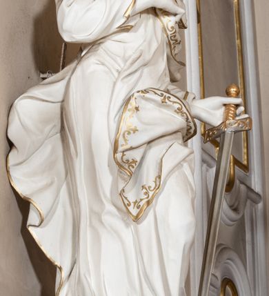 Zdjęcie nr 3: Rzeźba drążona z tyłu stoi na prostokątnej podstawie ustawionej na wolutowej konsoli przyściennej. Postać apostoła jest ujęta frontalnie w kontrapoście z głową skierowaną w prawo. Prawą rękę Paweł ma złożoną na piersi, lewą zaś wyciągniętą w bok trzyma miecz zwrócony ostrzem w dół. Święty ma pociągłą, wychudzoną twarz o migdałowatych oczach, lekko garbatym nosie i pełnych rozchylonych ustach. Głowę okala opadająca na pierś broda i lekko kręcone sięgające ramion włosy. Postać ubrana jest w długą, przylegającą do ciała suknię oraz płaszcz zarzucony na lewę ramię, opadający na plecy z fragmentem tkaniny przerzuconym przez lewe biodro. Rzeźba jest polichromowana w całości na biało ze złoconymi lamówkami szat i wzorem dekoracyjnym wzdłuż krawędzi. Miecz posiada złoconą rękojeść i srebrzony ostrze. 