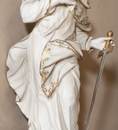 Zdjęcie nr 5: Rzeźba drążona z tyłu stoi na prostokątnej podstawie ustawionej na wolutowej konsoli przyściennej. Postać apostoła jest ujęta frontalnie w kontrapoście z głową skierowaną w prawo. Prawą rękę Paweł ma złożoną na piersi, lewą zaś wyciągniętą w bok trzyma miecz zwrócony ostrzem w dół. Święty ma pociągłą, wychudzoną twarz o migdałowatych oczach, lekko garbatym nosie i pełnych rozchylonych ustach. Głowę okala opadająca na pierś broda i lekko kręcone sięgające ramion włosy. Postać ubrana jest w długą, przylegającą do ciała suknię oraz płaszcz zarzucony na lewę ramię, opadający na plecy z fragmentem tkaniny przerzuconym przez lewe biodro. Rzeźba jest polichromowana w całości na biało ze złoconymi lamówkami szat i wzorem dekoracyjnym wzdłuż krawędzi. Miecz posiada złoconą rękojeść i srebrzony ostrze. 