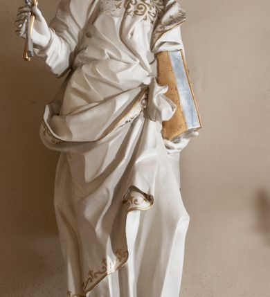 Zdjęcie nr 3: Rzeźba drążona z tyłu, stoi na prostokątnej podstawie ustawionej na wolutowej konsoli przyściennej. Postać apostoła jest ujęta frontalnie w kontrapoście z głową skierowaną i pochyloną w lewo. W prawej ręce odsuniętej od siebie apostoł trzyma klucze, a lewą podtrzymuje opartą o tors księgę. Piotr ma pociągłą, wychudzoną twarzą o migdałowatych oczach, małym nosie i pełnych ustach. Otacza ja krótka, kręcona broda i mocno kręcone włosy z łysiną na głowie. Postać ubrana jest w długą, przylegającą do ciała tunikę, przewiązaną w pasie oraz płaszcz zarzucony na lewe ramię, opadający na plecy, z fragmentem tkaniny przerzuconym przez prawe biodro, podwiązanym na wysokości pasa. Rzeźba została w całości polichromowana na biało ze złoconymi lamówkami szat i wzorem dekoracyjnym poprowadzonym wzdłuż krawędzi. Klucze i księga zostały wysrebrzone i wyzłocone.