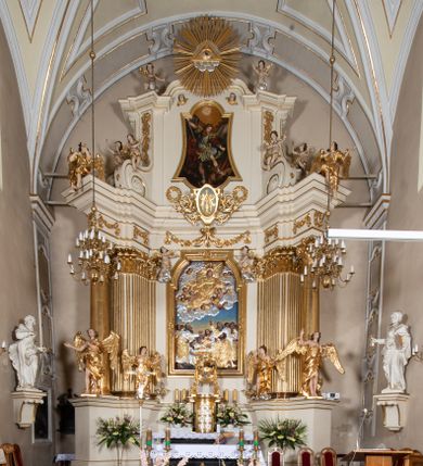 Zdjęcie nr 1: Dwukondygnacyjny, jednoosiowy ołtarz ze zwieńczeniem uzyskał strukturę architektoniczną. Stipes ołtarzowy wykonano w formie granitowego prostopadłościanu z mensą. Na mensie na osi ołtarza ustawiono tabernakulum z tronem wystawienia. Tabernakulum otrzymało kształt drewnianej skrzynki obitej blachą z drzwiczkami w formie stojącego prostokąta, z sześcioma kwaterami przedstawiającymi sceny z Pisma Świętego. Całość zamknięto silnie wysuniętym gzymsem wypukłym od frontu. Tron wystawienia utworzono w formie baldachimu ozdobionego lambrekinem z chwostami. Ujmują go spływy wolutowe z podwieszoną tkaniną, dekorowane liśćmi akantu. W przestrzeni zaplecka tronu umieszczono w promienistej glorii medalion z chrystogramem "IHS". Nastawa została ustawiona na wysokim cokole, zwieńczonym profilowanym gzymsem z wyodrębnionymi dwoma wspornikami skierowanymi do wewnątrz. Retabulum ujmuje  zwrócona na zewnątrz opilastrowana para filarów, przed którymi stoi para kolumn o kapitelach kompozytowych dźwigających pełne belkowanie wyłamane na ich osi. W polu centralnym znajduje się nisza w kształcie stojącego prostokąta zwieńczonego przerwanym prostokątnie łukiem półkolistym, nadwieszonym. Niszę obramienia profilowana rama. W jej wnętrzu znajduje się obraz Wniebowzięcia Najświętsze Marii Panny w drewnianych sukienkach wotywnych. Druga kondygnacja ujęta została dwiema parami zwróconych na zewnątrz pilastrów o trzonach przechodzących w spływy wolutowe. Pilastry dźwigają gzyms wyłamany na ich osi, który w części centralnej przyjmuje kształt spłaszczonego łuku. W polu umieszczono obraz z przedstawieniem Michała Archanioła w formie stojącego prostokąta o wklęsłych, dłuższych krawędziach oraz krótszych o kształcie łuku półkolistego nadwieszonego. Nastawa zakończona została okiem Opatrzności Bożej w glorii promienistej. Na cokole przed podporami ustawiono cztery figury adorujących aniołów. Ramę w polu środkowym udekorowano ornamentem roślinnym. W górnych przyłuczach umieszczono figury puttów na obłokach. Fryz został ozdobiony kwiatowymi girlandami. Centralnie na osi na gzymsie zawieszono kartusz z monogramem Marii w polu. Na zewnętrznych krawędziach belkowania pierwszej kondygnacji siedzą dwie figury aniołów, a na spływach wolutowych cztery putta. W trzonach pilastrów drugiej kondygnacji widoczne są zwisy z ornamentem kwiatowym podwieszonym na wstążce. W polu centralnym na dole umieszczono dwa wieńce, a u góry dwie uskrzydlone główki anielskie. Na belkowaniu znajdują się dwa putta na obłokach. Struktura nastawy została polichromowana w kolorze kości słoniowej, natomiast kolumny, pilastry, profile i ornamentyka zostały wyzłocone, a obłoki posrebrzone.