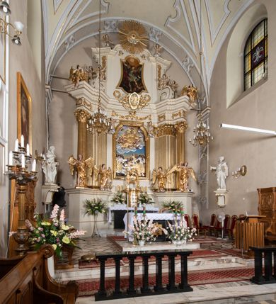 Zdjęcie nr 3: Dwukondygnacyjny, jednoosiowy ołtarz ze zwieńczeniem uzyskał strukturę architektoniczną. Stipes ołtarzowy wykonano w formie granitowego prostopadłościanu z mensą. Na mensie na osi ołtarza ustawiono tabernakulum z tronem wystawienia. Tabernakulum otrzymało kształt drewnianej skrzynki obitej blachą z drzwiczkami w formie stojącego prostokąta, z sześcioma kwaterami przedstawiającymi sceny z Pisma Świętego. Całość zamknięto silnie wysuniętym gzymsem wypukłym od frontu. Tron wystawienia utworzono w formie baldachimu ozdobionego lambrekinem z chwostami. Ujmują go spływy wolutowe z podwieszoną tkaniną, dekorowane liśćmi akantu. W przestrzeni zaplecka tronu umieszczono w promienistej glorii medalion z chrystogramem "IHS". Nastawa została ustawiona na wysokim cokole, zwieńczonym profilowanym gzymsem z wyodrębnionymi dwoma wspornikami skierowanymi do wewnątrz. Retabulum ujmuje  zwrócona na zewnątrz opilastrowana para filarów, przed którymi stoi para kolumn o kapitelach kompozytowych dźwigających pełne belkowanie wyłamane na ich osi. W polu centralnym znajduje się nisza w kształcie stojącego prostokąta zwieńczonego przerwanym prostokątnie łukiem półkolistym, nadwieszonym. Niszę obramienia profilowana rama. W jej wnętrzu znajduje się obraz Wniebowzięcia Najświętsze Marii Panny w drewnianych sukienkach wotywnych. Druga kondygnacja ujęta została dwiema parami zwróconych na zewnątrz pilastrów o trzonach przechodzących w spływy wolutowe. Pilastry dźwigają gzyms wyłamany na ich osi, który w części centralnej przyjmuje kształt spłaszczonego łuku. W polu umieszczono obraz z przedstawieniem Michała Archanioła w formie stojącego prostokąta o wklęsłych, dłuższych krawędziach oraz krótszych o kształcie łuku półkolistego nadwieszonego. Nastawa zakończona została okiem Opatrzności Bożej w glorii promienistej. Na cokole przed podporami ustawiono cztery figury adorujących aniołów. Ramę w polu środkowym udekorowano ornamentem roślinnym. W górnych przyłuczach umieszczono figury puttów na obłokach. Fryz został ozdobiony kwiatowymi girlandami. Centralnie na osi na gzymsie zawieszono kartusz z monogramem Marii w polu. Na zewnętrznych krawędziach belkowania pierwszej kondygnacji siedzą dwie figury aniołów, a na spływach wolutowych cztery putta. W trzonach pilastrów drugiej kondygnacji widoczne są zwisy z ornamentem kwiatowym podwieszonym na wstążce. W polu centralnym na dole umieszczono dwa wieńce, a u góry dwie uskrzydlone główki anielskie. Na belkowaniu znajdują się dwa putta na obłokach. Struktura nastawy została polichromowana w kolorze kości słoniowej, natomiast kolumny, pilastry, profile i ornamentyka zostały wyzłocone, a obłoki posrebrzone.