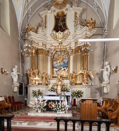 Zdjęcie nr 2: Dwukondygnacyjny, jednoosiowy ołtarz ze zwieńczeniem uzyskał strukturę architektoniczną. Stipes ołtarzowy wykonano w formie granitowego prostopadłościanu z mensą. Na mensie na osi ołtarza ustawiono tabernakulum z tronem wystawienia. Tabernakulum otrzymało kształt drewnianej skrzynki obitej blachą z drzwiczkami w formie stojącego prostokąta, z sześcioma kwaterami przedstawiającymi sceny z Pisma Świętego. Całość zamknięto silnie wysuniętym gzymsem wypukłym od frontu. Tron wystawienia utworzono w formie baldachimu ozdobionego lambrekinem z chwostami. Ujmują go spływy wolutowe z podwieszoną tkaniną, dekorowane liśćmi akantu. W przestrzeni zaplecka tronu umieszczono w promienistej glorii medalion z chrystogramem "IHS". Nastawa została ustawiona na wysokim cokole, zwieńczonym profilowanym gzymsem z wyodrębnionymi dwoma wspornikami skierowanymi do wewnątrz. Retabulum ujmuje  zwrócona na zewnątrz opilastrowana para filarów, przed którymi stoi para kolumn o kapitelach kompozytowych dźwigających pełne belkowanie wyłamane na ich osi. W polu centralnym znajduje się nisza w kształcie stojącego prostokąta zwieńczonego przerwanym prostokątnie łukiem półkolistym, nadwieszonym. Niszę obramienia profilowana rama. W jej wnętrzu znajduje się obraz Wniebowzięcia Najświętsze Marii Panny w drewnianych sukienkach wotywnych. Druga kondygnacja ujęta została dwiema parami zwróconych na zewnątrz pilastrów o trzonach przechodzących w spływy wolutowe. Pilastry dźwigają gzyms wyłamany na ich osi, który w części centralnej przyjmuje kształt spłaszczonego łuku. W polu umieszczono obraz z przedstawieniem Michała Archanioła w formie stojącego prostokąta o wklęsłych, dłuższych krawędziach oraz krótszych o kształcie łuku półkolistego nadwieszonego. Nastawa zakończona została okiem Opatrzności Bożej w glorii promienistej. Na cokole przed podporami ustawiono cztery figury adorujących aniołów. Ramę w polu środkowym udekorowano ornamentem roślinnym. W górnych przyłuczach umieszczono figury puttów na obłokach. Fryz został ozdobiony kwiatowymi girlandami. Centralnie na osi na gzymsie zawieszono kartusz z monogramem Marii w polu. Na zewnętrznych krawędziach belkowania pierwszej kondygnacji siedzą dwie figury aniołów, a na spływach wolutowych cztery putta. W trzonach pilastrów drugiej kondygnacji widoczne są zwisy z ornamentem kwiatowym podwieszonym na wstążce. W polu centralnym na dole umieszczono dwa wieńce, a u góry dwie uskrzydlone główki anielskie. Na belkowaniu znajdują się dwa putta na obłokach. Struktura nastawy została polichromowana w kolorze kości słoniowej, natomiast kolumny, pilastry, profile i ornamentyka zostały wyzłocone, a obłoki posrebrzone.