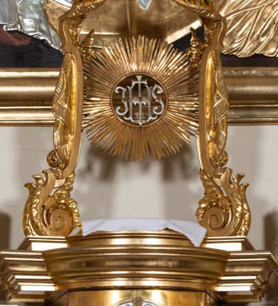 Zdjęcie nr 9: Dwukondygnacyjny, jednoosiowy ołtarz ze zwieńczeniem uzyskał strukturę architektoniczną. Stipes ołtarzowy wykonano w formie granitowego prostopadłościanu z mensą. Na mensie na osi ołtarza ustawiono tabernakulum z tronem wystawienia. Tabernakulum otrzymało kształt drewnianej skrzynki obitej blachą z drzwiczkami w formie stojącego prostokąta, z sześcioma kwaterami przedstawiającymi sceny z Pisma Świętego. Całość zamknięto silnie wysuniętym gzymsem wypukłym od frontu. Tron wystawienia utworzono w formie baldachimu ozdobionego lambrekinem z chwostami. Ujmują go spływy wolutowe z podwieszoną tkaniną, dekorowane liśćmi akantu. W przestrzeni zaplecka tronu umieszczono w promienistej glorii medalion z chrystogramem &quot;IHS&quot;. Nastawa została ustawiona na wysokim cokole, zwieńczonym profilowanym gzymsem z wyodrębnionymi dwoma wspornikami skierowanymi do wewnątrz. Retabulum ujmuje  zwrócona na zewnątrz opilastrowana para filarów, przed którymi stoi para kolumn o kapitelach kompozytowych dźwigających pełne belkowanie wyłamane na ich osi. W polu centralnym znajduje się nisza w kształcie stojącego prostokąta zwieńczonego przerwanym prostokątnie łukiem półkolistym, nadwieszonym. Niszę obramienia profilowana rama. W jej wnętrzu znajduje się obraz Wniebowzięcia Najświętsze Marii Panny w drewnianych sukienkach wotywnych. Druga kondygnacja ujęta została dwiema parami zwróconych na zewnątrz pilastrów o trzonach przechodzących w spływy wolutowe. Pilastry dźwigają gzyms wyłamany na ich osi, który w części centralnej przyjmuje kształt spłaszczonego łuku. W polu umieszczono obraz z przedstawieniem Michała Archanioła w formie stojącego prostokąta o wklęsłych, dłuższych krawędziach oraz krótszych o kształcie łuku półkolistego nadwieszonego. Nastawa zakończona została okiem Opatrzności Bożej w glorii promienistej. Na cokole przed podporami ustawiono cztery figury adorujących aniołów. Ramę w polu środkowym udekorowano ornamentem roślinnym. W górnych przyłuczach umieszczono figury puttów na obłokach. Fryz został ozdobiony kwiatowymi girlandami. Centralnie na osi na gzymsie zawieszono kartusz z monogramem Marii w polu. Na zewnętrznych krawędziach belkowania pierwszej kondygnacji siedzą dwie figury aniołów, a na spływach wolutowych cztery putta. W trzonach pilastrów drugiej kondygnacji widoczne są zwisy z ornamentem kwiatowym podwieszonym na wstążce. W polu centralnym na dole umieszczono dwa wieńce, a u góry dwie uskrzydlone główki anielskie. Na belkowaniu znajdują się dwa putta na obłokach. Struktura nastawy została polichromowana w kolorze kości słoniowej, natomiast kolumny, pilastry, profile i ornamentyka zostały wyzłocone, a obłoki posrebrzone.