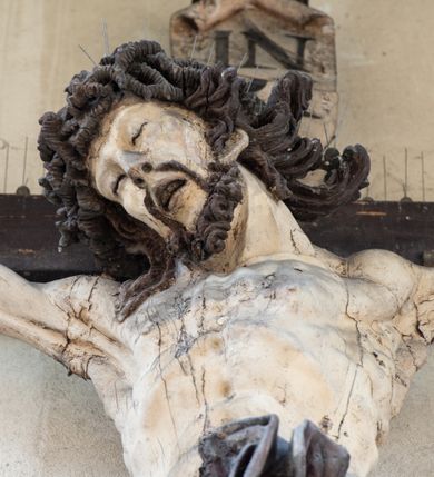 Zdjęcie nr 4: Figura Chrystusa umarłego została przybita do drewnianego krzyża za pomocą trzech gwoździ. Ciało Zbawiciela ujęto w lekkim zwisie o szeroko rozłożonych ramionach. Jego głowa bezwładnie opada na prawy bark, nogi zostały ugięte w kolanach, stopy skrzyżowane w układzie prawa na lewą.  Twarz Jezusa jest szczupła o zamkniętych oczach, prostym nosie i otwartych ustach. Okala ją krótka, silnie kręcona broda oraz długie, skręcające się w pukle i rozwiane włosy. Na głowie widoczna jest korona cierniowa. Ciało Chrystusa jest wychudzone z silnie podkreślonymi wklęsło-wypukłymi mięśniami i zaznaczonymi żebrami. Perizonium widoczne na biodrach zostało zawieszone na podwójnym sznurze odsłaniającym lewe biodro z silnie drapowaną i ekspresyjnie rozwianą tkaniną. Ciało zostało polichromowane w kolorze naturalnym, włosy i perizonium są brązowe.