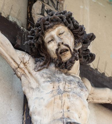 Zdjęcie nr 3: Figura Chrystusa umarłego została przybita do drewnianego krzyża za pomocą trzech gwoździ. Ciało Zbawiciela ujęto w lekkim zwisie o szeroko rozłożonych ramionach. Jego głowa bezwładnie opada na prawy bark, nogi zostały ugięte w kolanach, stopy skrzyżowane w układzie prawa na lewą.  Twarz Jezusa jest szczupła o zamkniętych oczach, prostym nosie i otwartych ustach. Okala ją krótka, silnie kręcona broda oraz długie, skręcające się w pukle i rozwiane włosy. Na głowie widoczna jest korona cierniowa. Ciało Chrystusa jest wychudzone z silnie podkreślonymi wklęsło-wypukłymi mięśniami i zaznaczonymi żebrami. Perizonium widoczne na biodrach zostało zawieszone na podwójnym sznurze odsłaniającym lewe biodro z silnie drapowaną i ekspresyjnie rozwianą tkaniną. Ciało zostało polichromowane w kolorze naturalnym, włosy i perizonium są brązowe.