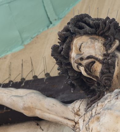 Zdjęcie nr 5: Figura Chrystusa umarłego została przybita do drewnianego krzyża za pomocą trzech gwoździ. Ciało Zbawiciela ujęto w lekkim zwisie o szeroko rozłożonych ramionach. Jego głowa bezwładnie opada na prawy bark, nogi zostały ugięte w kolanach, stopy skrzyżowane w układzie prawa na lewą.  Twarz Jezusa jest szczupła o zamkniętych oczach, prostym nosie i otwartych ustach. Okala ją krótka, silnie kręcona broda oraz długie, skręcające się w pukle i rozwiane włosy. Na głowie widoczna jest korona cierniowa. Ciało Chrystusa jest wychudzone z silnie podkreślonymi wklęsło-wypukłymi mięśniami i zaznaczonymi żebrami. Perizonium widoczne na biodrach zostało zawieszone na podwójnym sznurze odsłaniającym lewe biodro z silnie drapowaną i ekspresyjnie rozwianą tkaniną. Ciało zostało polichromowane w kolorze naturalnym, włosy i perizonium są brązowe.