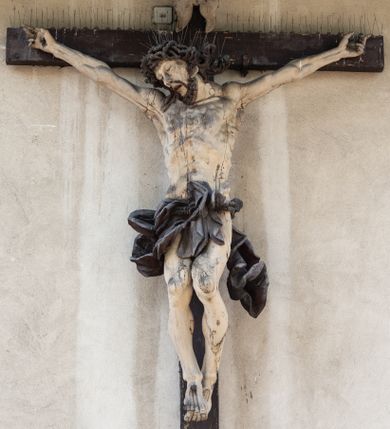 Zdjęcie nr 1: Figura Chrystusa umarłego została przybita do drewnianego krzyża za pomocą trzech gwoździ. Ciało Zbawiciela ujęto w lekkim zwisie o szeroko rozłożonych ramionach. Jego głowa bezwładnie opada na prawy bark, nogi zostały ugięte w kolanach, stopy skrzyżowane w układzie prawa na lewą.  Twarz Jezusa jest szczupła o zamkniętych oczach, prostym nosie i otwartych ustach. Okala ją krótka, silnie kręcona broda oraz długie, skręcające się w pukle i rozwiane włosy. Na głowie widoczna jest korona cierniowa. Ciało Chrystusa jest wychudzone z silnie podkreślonymi wklęsło-wypukłymi mięśniami i zaznaczonymi żebrami. Perizonium widoczne na biodrach zostało zawieszone na podwójnym sznurze odsłaniającym lewe biodro z silnie drapowaną i ekspresyjnie rozwianą tkaniną. Ciało zostało polichromowane w kolorze naturalnym, włosy i perizonium są brązowe.