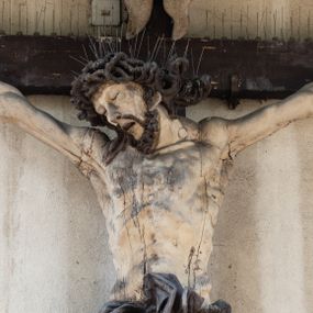 Zdjęcie nr 1: Figura Chrystusa umarłego została przybita do drewnianego krzyża za pomocą trzech gwoździ. Ciało Zbawiciela ujęto w lekkim zwisie o szeroko rozłożonych ramionach. Jego głowa bezwładnie opada na prawy bark, nogi zostały ugięte w kolanach, stopy skrzyżowane w układzie prawa na lewą.  Twarz Jezusa jest szczupła o zamkniętych oczach, prostym nosie i otwartych ustach. Okala ją krótka, silnie kręcona broda oraz długie, skręcające się w pukle i rozwiane włosy. Na głowie widoczna jest korona cierniowa. Ciało Chrystusa jest wychudzone z silnie podkreślonymi wklęsło-wypukłymi mięśniami i zaznaczonymi żebrami. Perizonium widoczne na biodrach zostało zawieszone na podwójnym sznurze odsłaniającym lewe biodro z silnie drapowaną i ekspresyjnie rozwianą tkaniną. Ciało zostało polichromowane w kolorze naturalnym, włosy i perizonium są brązowe.