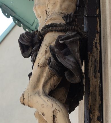 Zdjęcie nr 7: Figura Chrystusa umarłego została przybita do drewnianego krzyża za pomocą trzech gwoździ. Ciało Zbawiciela ujęto w lekkim zwisie o szeroko rozłożonych ramionach. Jego głowa bezwładnie opada na prawy bark, nogi zostały ugięte w kolanach, stopy skrzyżowane w układzie prawa na lewą.  Twarz Jezusa jest szczupła o zamkniętych oczach, prostym nosie i otwartych ustach. Okala ją krótka, silnie kręcona broda oraz długie, skręcające się w pukle i rozwiane włosy. Na głowie widoczna jest korona cierniowa. Ciało Chrystusa jest wychudzone z silnie podkreślonymi wklęsło-wypukłymi mięśniami i zaznaczonymi żebrami. Perizonium widoczne na biodrach zostało zawieszone na podwójnym sznurze odsłaniającym lewe biodro z silnie drapowaną i ekspresyjnie rozwianą tkaniną. Ciało zostało polichromowane w kolorze naturalnym, włosy i perizonium są brązowe.