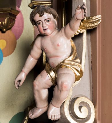 Zdjęcie nr 5: Przyścienna, wisząca ambona składa się z kosza, zaplecka i baldachimu. Wnętrze ambony dostępne jest przez drzwiczki umieszczone w polu zaplecka za pomocą schodów prowadzących z zakrystii. Kosz wykonano na planie prostokąta o wypukłej dłuższej ściance i wklęsłych krótszych, ustawiony na spłaszczonym wałku. Jej spód zdobi podwieszenie w formie zbliżonej do stożka, ujętego czterema wolutami, zakończone elementem wolutowym. W narożach kosza znalazły się cztery pilastry o trzonach przechodzących w spływy wolutowe, dźwigających imposty oraz parapet w formie gzymsu. Na frontowych wolutach umieszczono siedzące putta. Ścianki zostały dekorowane prostokątnymi płycinami. Na ściance frontowej wyodrębniono płycinę o wklęsło wypukłym zarysie z umieszczonymi w polu dwiema uskrzydlonymi główkami anielskimi oraz ornamentem roślinnym. Zaplecek uzyskał formę stojącego prostokąta ujętego parą pilastrów z konsolami dźwigającymi imposty oraz profilowany gzyms. W polu zamontowano drzwiczki dekorowane płycinami z uskrzydlonymi główkami anielskimi. Baldachim powstał w formie podstawy na planie analogicznym względem kosza, obwiedziony profilowanym gzymsem z fragmentami przerwanego wolutowego przyczółka, na którym siedzą putta. Całość zwieńczono medalionem w otoku z obłoków ze srebrzoną gołębicą Ducha Świętego i trzema główkami anielskimi w glorii promienistej. Struktura została polichromowana w kolorze pudrowego różu z jasnofioletowymi detalami, o złoconych profilach i glorii.