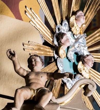 Zdjęcie nr 3: Przyścienna, wisząca ambona składa się z kosza, zaplecka i baldachimu. Wnętrze ambony dostępne jest przez drzwiczki umieszczone w polu zaplecka za pomocą schodów prowadzących z zakrystii. Kosz wykonano na planie prostokąta o wypukłej dłuższej ściance i wklęsłych krótszych, ustawiony na spłaszczonym wałku. Jej spód zdobi podwieszenie w formie zbliżonej do stożka, ujętego czterema wolutami, zakończone elementem wolutowym. W narożach kosza znalazły się cztery pilastry o trzonach przechodzących w spływy wolutowe, dźwigających imposty oraz parapet w formie gzymsu. Na frontowych wolutach umieszczono siedzące putta. Ścianki zostały dekorowane prostokątnymi płycinami. Na ściance frontowej wyodrębniono płycinę o wklęsło wypukłym zarysie z umieszczonymi w polu dwiema uskrzydlonymi główkami anielskimi oraz ornamentem roślinnym. Zaplecek uzyskał formę stojącego prostokąta ujętego parą pilastrów z konsolami dźwigającymi imposty oraz profilowany gzyms. W polu zamontowano drzwiczki dekorowane płycinami z uskrzydlonymi główkami anielskimi. Baldachim powstał w formie podstawy na planie analogicznym względem kosza, obwiedziony profilowanym gzymsem z fragmentami przerwanego wolutowego przyczółka, na którym siedzą putta. Całość zwieńczono medalionem w otoku z obłoków ze srebrzoną gołębicą Ducha Świętego i trzema główkami anielskimi w glorii promienistej. Struktura została polichromowana w kolorze pudrowego różu z jasnofioletowymi detalami, o złoconych profilach i glorii.