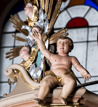 Zdjęcie nr 4: Przyścienna, wisząca ambona składa się z kosza, zaplecka i baldachimu. Wnętrze ambony dostępne jest przez drzwiczki umieszczone w polu zaplecka za pomocą schodów prowadzących z zakrystii. Kosz wykonano na planie prostokąta o wypukłej dłuższej ściance i wklęsłych krótszych, ustawiony na spłaszczonym wałku. Jej spód zdobi podwieszenie w formie zbliżonej do stożka, ujętego czterema wolutami, zakończone elementem wolutowym. W narożach kosza znalazły się cztery pilastry o trzonach przechodzących w spływy wolutowe, dźwigających imposty oraz parapet w formie gzymsu. Na frontowych wolutach umieszczono siedzące putta. Ścianki zostały dekorowane prostokątnymi płycinami. Na ściance frontowej wyodrębniono płycinę o wklęsło wypukłym zarysie z umieszczonymi w polu dwiema uskrzydlonymi główkami anielskimi oraz ornamentem roślinnym. Zaplecek uzyskał formę stojącego prostokąta ujętego parą pilastrów z konsolami dźwigającymi imposty oraz profilowany gzyms. W polu zamontowano drzwiczki dekorowane płycinami z uskrzydlonymi główkami anielskimi. Baldachim powstał w formie podstawy na planie analogicznym względem kosza, obwiedziony profilowanym gzymsem z fragmentami przerwanego wolutowego przyczółka, na którym siedzą putta. Całość zwieńczono medalionem w otoku z obłoków ze srebrzoną gołębicą Ducha Świętego i trzema główkami anielskimi w glorii promienistej. Struktura została polichromowana w kolorze pudrowego różu z jasnofioletowymi detalami, o złoconych profilach i glorii.